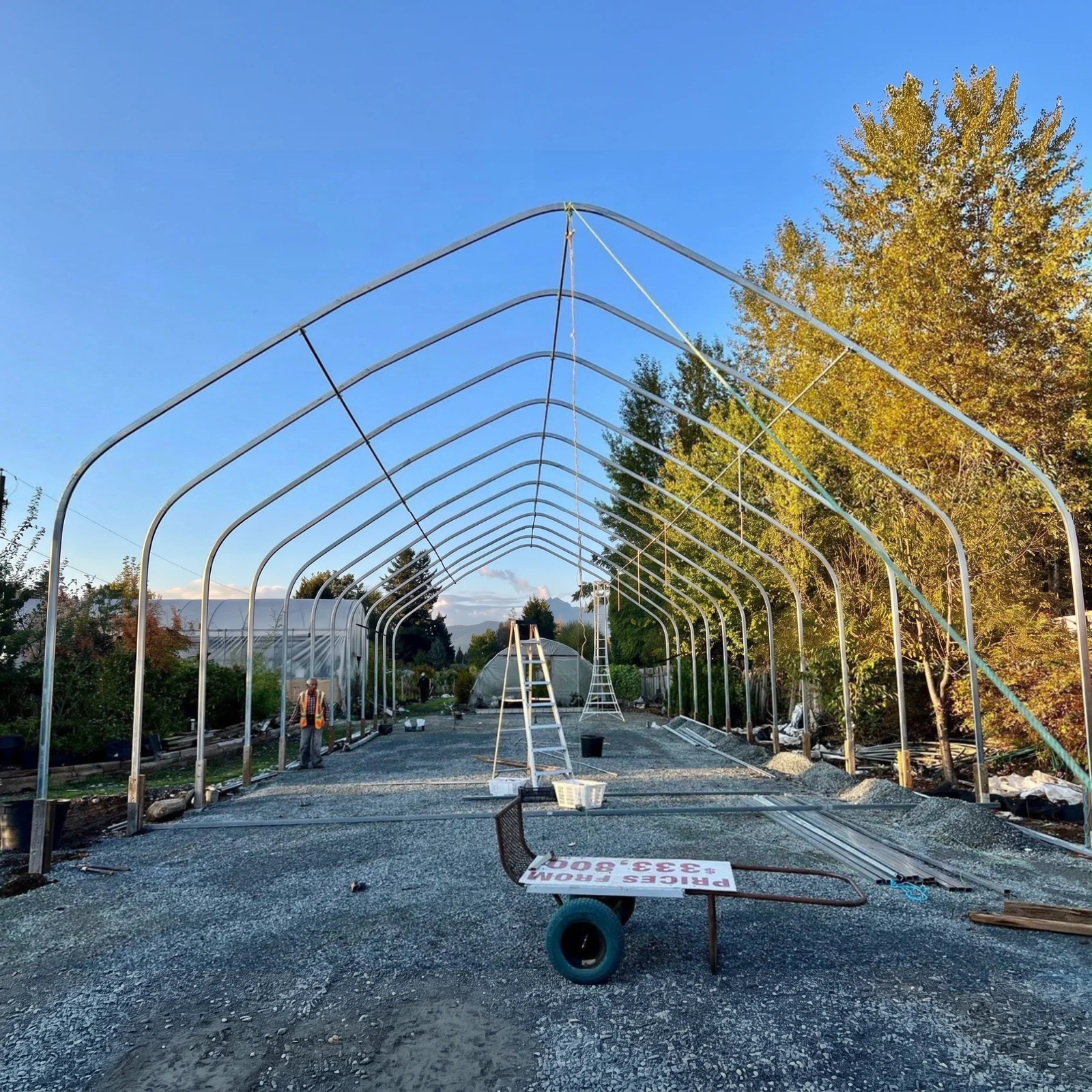 Sun & Steel high tunnel frame under construction in Maple Ridge, BC — galvanized arches raised on posts above grade for extra interior height, on a fresh gravel pad